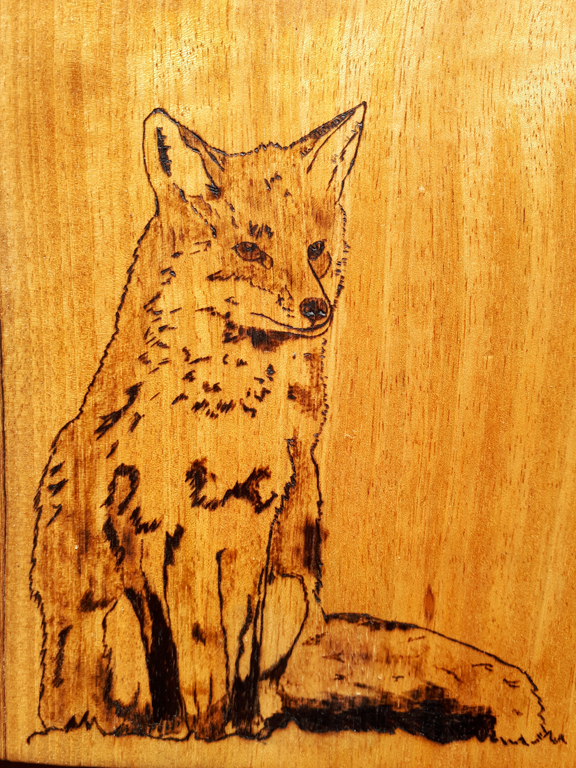 Light wood, vertical grain. the fox is sitting straight up with its tail sticking out to the left. It's looking off into the distance. Monochromatic brown, the design is burnt into the wood.