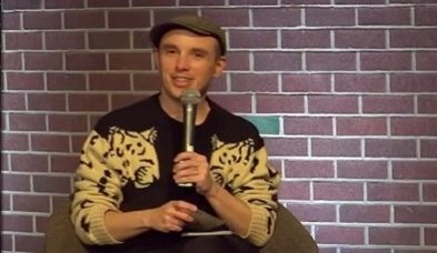 AJ Withers holding microphone and wearing the best sweater ever (white lepoards as the arms with their heads on the shoulders, blak body), siting in front of a brick wall.